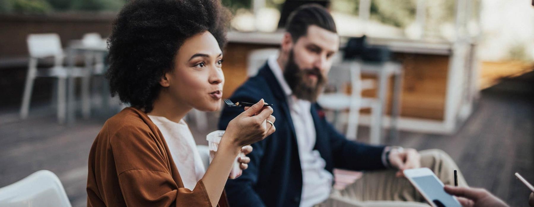 woman sits and eats outside while someone holding a cell talks to her